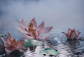 Fotobehang Happy young little blonde woman sits in a huge pink lotus flower that floats on the water. Fantasy artistic photo Beautiful girl in a fairy tale Thumbelina. Goddess of nature on the river in blue fog © kharchenkoirina