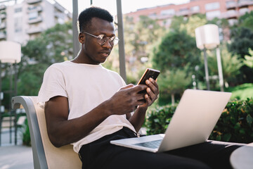 Concentrated african american hipster guy in eyewear for vision correction reading notification on smartphone, serious dark skinned businessman checking mails on mobile phone on remote job