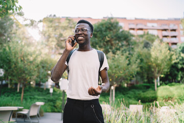 Cheerful dark skinned hipster guy in eyewear satisfied with roaming tariffs and connection walking on urban settings, happy african american male having positive mobile phone conversation