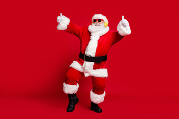 Full body size photo of pensioner old man grey beard show thumbs up party dancing wear santa x-mas costume coat sunglass headwear wireless earphones boots isolated red color background