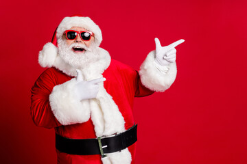 Photo of retired old man grey beard open mouth smile direct fingers empty space demonstrate best option wear santa x-mas costume gloves coat sunglass headwear isolated red color background