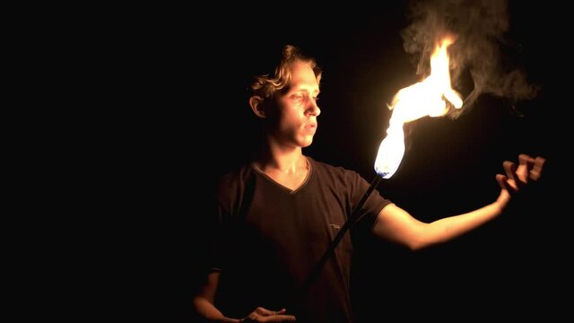 Young guy playing with fire. He brings the burning torch to him and passes his hand over it. Dangerous stunt. Fire show