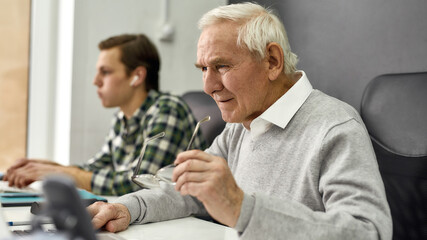 Close up of aged man, senior intern looking focused while using laptop, sitting at desk, working in modern office with young colleague