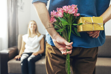 husband hiding romantic surprise present and flowers behind back to beloved wife at home
