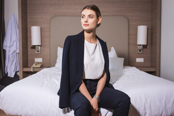 young businesswoman in suit looking away and sitting in bed on hotel