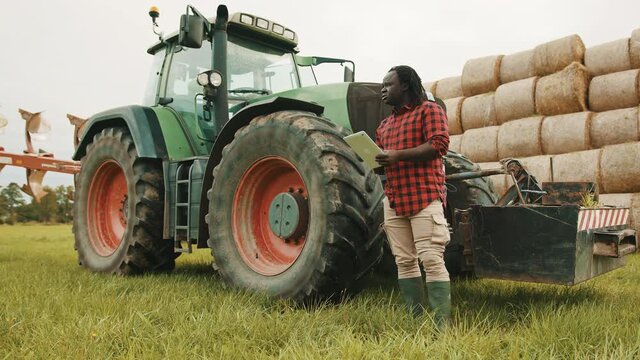 Young african farmer using tablet in front og big green tractor anf haystack. High quality 4k footage