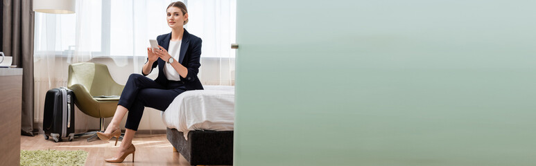 horizontal image of young businesswoman in suit holding smartphone while sitting on bed near travel bag in hotel room