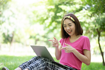 Asian woman pointed hands to laptop screen for hello her friends in video call and smiling face. Relax and comfortable in the public park. Education concept.