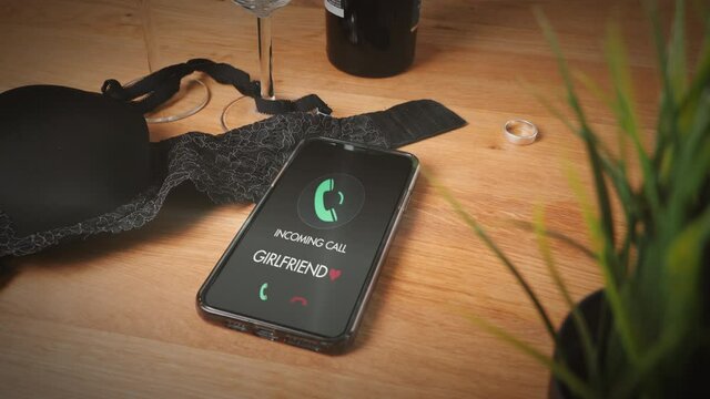 Concept affair with incoming phone call from girlfriend. Brassiere and wedding ring are on table. Husband liaison. Close up focus of smartphone. Girlfriend is calling. Cheating, obsession, possession.