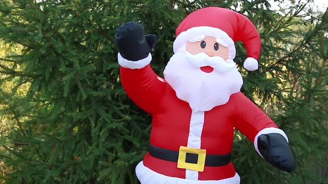A life-size inflatable Santa Claus doll in a red suit stands swaying, waving against a background of green trees. Happy New year celebration, happy holiday