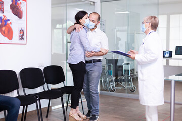 Disappointed young couple in hospital waiting area from doctor unfavorable bad news during coronavirus outbreak. Medic wearing face mask against infection with covid-19.