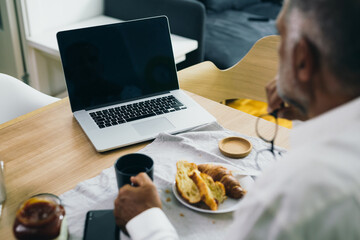 mid aged man having breakfast at home and using laptop computer