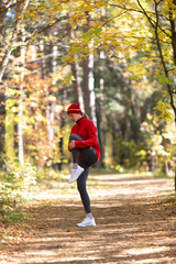 Caucasian fit woman in red sportswear and a cap warning up her knees before jogging in the forest in autumn, selective focus