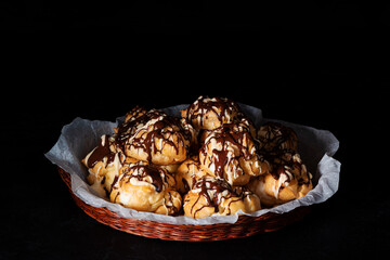 Homemade profiteroles with custard cream topped with melted dark and white chocolate in wicker basket on black background. Traditional French dessert. Low key. Copy space.