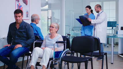 Fototapeta premium Nurse looking at radiography while talking with doctor about patient diagnosis in hospital waiting area. Invalid old woman in wheelchair. Senior man waiting for medical examination.