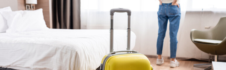 selective focus of yellow luggage near woman standing in hotel room, horizontal crop - obrazy, fototapety, plakaty