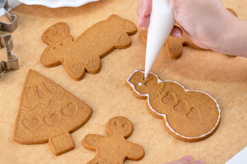 Young woman is decorating Christmas Gingerbread House cookies biscuit at home with frosting topping in icing bag, close up, lifestyle.
