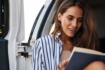 Beautiful young woman reading book while sitting in a back of her car outdoors