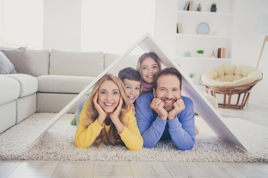 Safe family new house concept. Photo of people mom dad two small kids lying floor under paper card roof in living room indoors