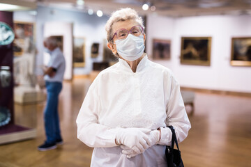 Fototapeta na wymiar elderly European woman in mask protecting against covid examines paintings on display in hall of art museum