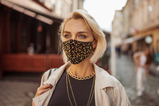 Outdoor fashion, lifestyle portrait of elegant woman wearing trendy outfit with protective face mask, many golden chain necklaces, walking in street of European city. Copy, empty space for text