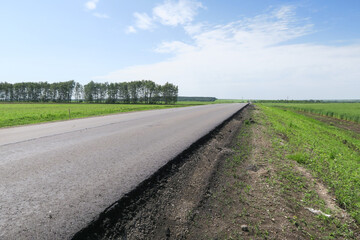 Fototapeta na wymiar New asphalt road through the field. Close-up of an empty asphalt road. Clear horizon, blue sky and low-slung road. Green grass and yellow sunflowers on the roadside.