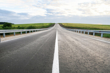 Obraz premium New asphalt road through the field. Close-up of an empty asphalt road. Clear horizon, blue sky and low-slung road. Green grass and yellow sunflowers on the roadside.