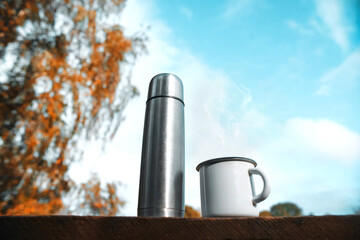 Thermos with a mug on a background of blue sky. Close-up. The concept of a picnic in nature, travel to the wild. Autumn forest, yellow foliage