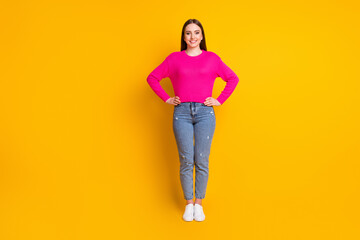 Full length photo of lovely young woman hands hips look camera wear pink sweater jeans sneakers isolated yellow color background