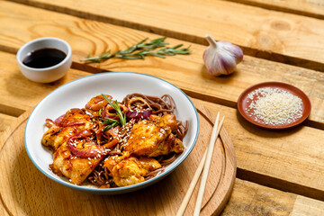 Soby with chicken and sesame seeds with onions in a gray plate on a wooden stand and sticks next to sesame seeds, and soy sauce on a wooden table.