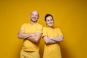Funny, cute man and woman in plain bright clothes are standing with crossed arms and smiling benevolent, on a yellow background.