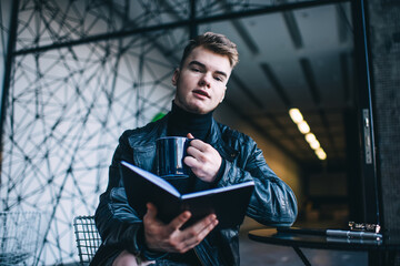 Young male drinking coffee and reading book in modern cafe