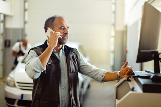 Happy mechanic talking on the phone while using computer at auto repair shop.