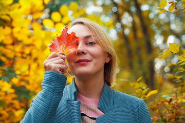 Smiling blonde woman covering one eye with red maple leaf. Autumn concept. Outdoor fall female portrait close-up