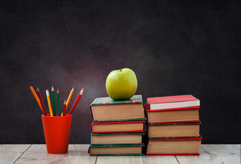 pile of books in colorful covers, pencils in holder and green apple on wooden table with green blackboard background. Distance home education. Back to school, quarantine concept of stay home.