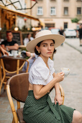 beautiful stylish young woman in a hat sitting at a table in a cafe and drinking coffee