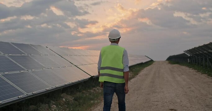 Backside view of male engineer in uniform walking and looking at solar power plant. Man in hard helmet examining object. Concept of solar station development and green energy.