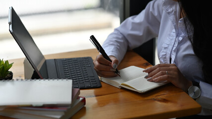 Closeup female freelancer writing her project on notebook and using laptop on wooden desk.