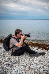 A caucasian male tourist photographer sitting on pebbles stone beach by the water with horizon in the background on the island Gotland in the Baltic Sea.