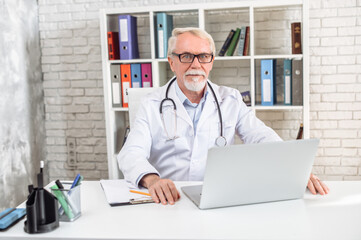 Mature gray haired male doctor is using laptop in hospital office