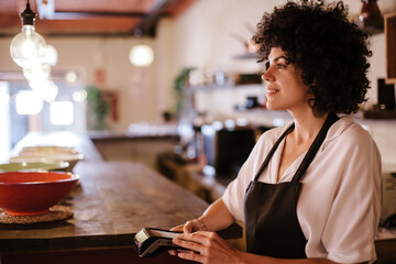 Portrait of a caucasian female barista working with the card terminal. Woman with apron standing behind the bar counter of a restaurant. Concept small business owner, young entrepreneur.