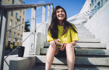 Happy asian woman in casual wear enjoying spending free time on urban setting during sunny day, smiling hipster girl 20s laughing and having fun in city sitting on stairs during summer leisure