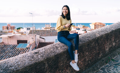 Portrait of cheerful Asian woman dressed in casual clothing smiling at camera during travel vacations, happy female tourist with modern cellphone device in hand posing at overlooking street area