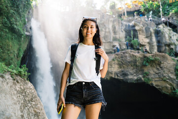 Cheerful female traveler with phone standing against waterfall