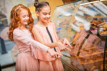 Little girl friends stand at shop window in a cafe and choose buns and cakes.