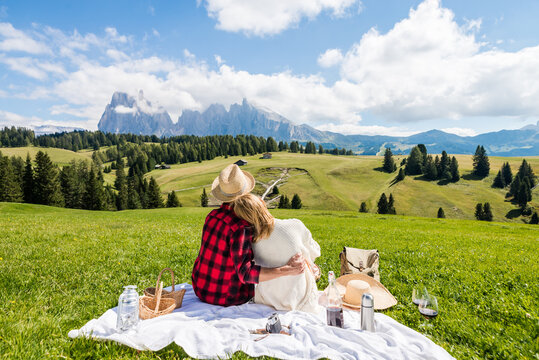 Young couple in love doing picnic visiting alps dolomities. Boyfriend and girlfriend sitting and looking at the beautiful scenic green meadow landscape.