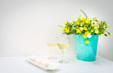 Romantic set up of burning candles, two  white wine glasses and blooming flowers in bucket on white table and wall in background