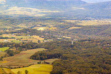 A scenic vista of Shenandoah Valley as seen from a scenic overlook by Skyline drive. There are small farms within a vast forest on valley floor with buildings and farmhouses nearby.
