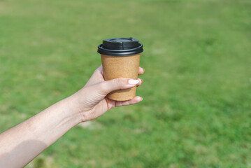 left hand of a caucasian young woman holding a paper cup of take away coffee in a green park