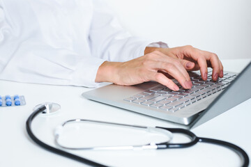 hands of a caucasian female doctor typing on a modern laptop on the top of a white desk